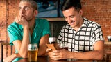 Zwei Männer sitzen bei einem Bier zusammen, der eine guckt nur auf sein Handy, der andere langweilt sich.