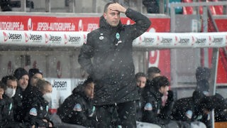 Werder-Trainer Ole Werner steht bei leichtem Schneefall in Regensburg an der Seitenlinie und fasst sich ungläubig mit der Hand an die Stirn.