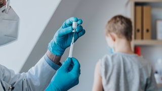 Ein Arzt zieht eine Impfdosis auf eine Spritze auf. Im Hintergrund sieht man einen Jungen sitzen.