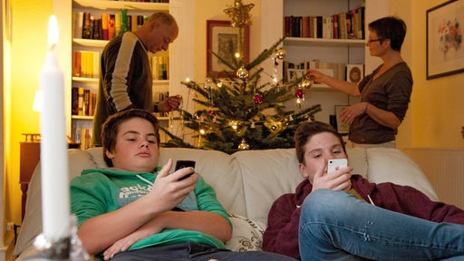 Zwei Jugendliche in Ingelheim am Rhein spielen mit ihren Handy, im Hintergrund die Eltern, die den Weihnachtsbaum schmücken