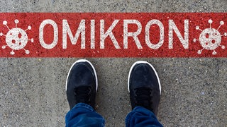 Zwei Füße stoppen vor der Aufschrift "Omikron" auf dem Asphalt