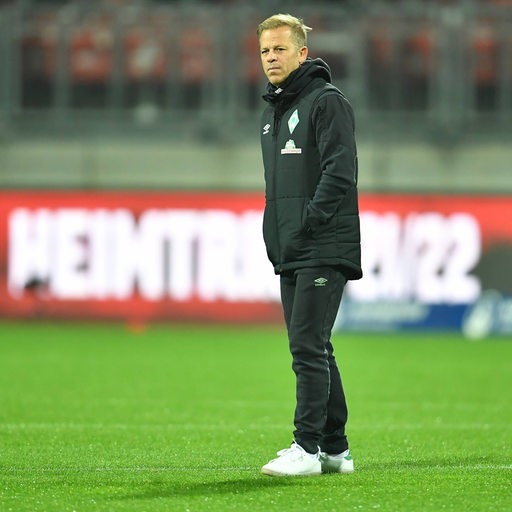 Werders ehemaliger Trainer Markus Anfang steht, die Hände in den Jackentaschen, auf einem Fußballplatz.
