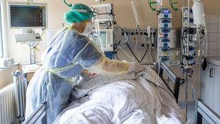 Eine Krankenschwester in Schutzkleidung beugt sich über einen Patienten, der in einem Bett liegt und an Geräte angeschlossen ist.