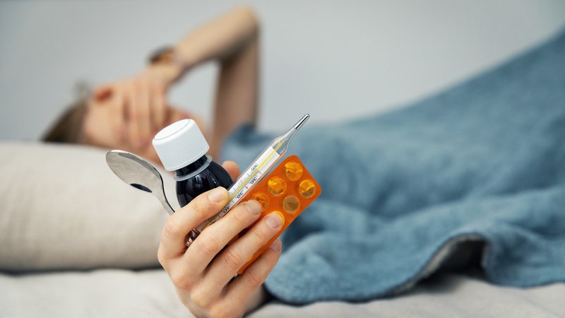 Kranke Frau im Bett, im Vordergrund hält sie Medizin und ein Fieberthermometer in der Hand