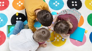 Drei Kinder spielen mit Tierfiguren auf einem bunten Teppich.