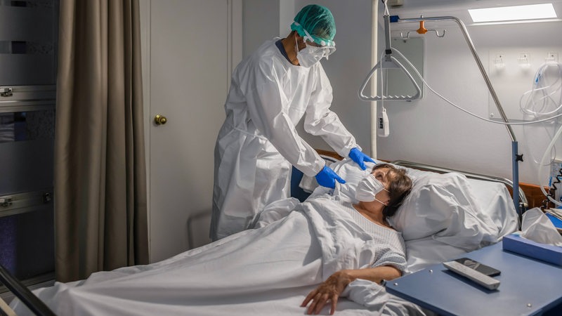 Ein Mann in Schutzkleidung versorgt eine Frau, die in einem Krankenhausbett liegt.