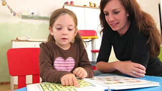 Ein Mädchen im Kindergartenalter zeigt neben einer Erzieherin in einem Bilderbuch auf ein Bild