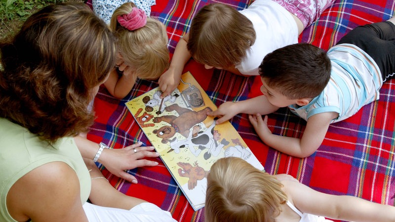 Sprachförderung in der Kita. Kinder liegen auf einer Decke und zeigen auf Bilder in einem Bilderbuch. Die Erzieherin sitzt daneben