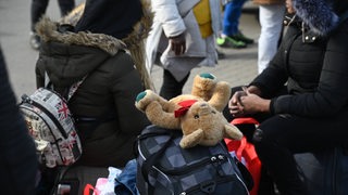 Ein Mädchen und eine Frau sitzen neben Koffern und Taschen. Auf der obersten Tasche liegt ein Teddy.