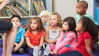 Eine Grupper Kinder sitzt auf dem Boden und hört einer Erwachsenen zu.