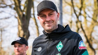 Trainer Ole Werner mit breitem Lächeln und schwarzer Kappe auf dem Weg zum Werder-Training.