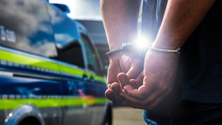 Ein Mann steht mit Handschellen vor einem Polizeiwagen.
