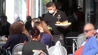 In einer gut besetzten Außen-Gastronomie serviert ein Kellner mit Mund-Nasenschutz einen Burger mit Beilagen.