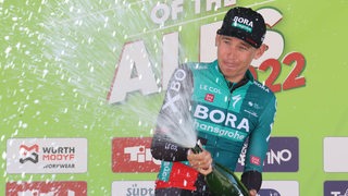 Radprofi Lennard Kämna feiert auf dem Podest seinen Etappensieg bei der Tour of the Alps mit einer Sektdusche.
