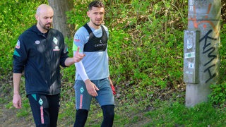 Werder-Verteidiger Milos Veljkovic neben dem Fitness-Coach auf dem Weg in die Kabine, nachdem er das Training abbrechen musste.