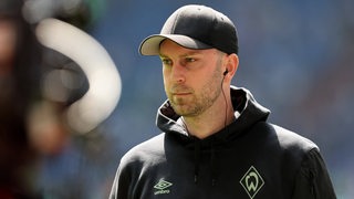 Werder-Trainer Ole Werner mit schwarzer Baseball-Kappe und Ohrhörern bei einem Interview vor dem Spielbeginn auf Schalke.