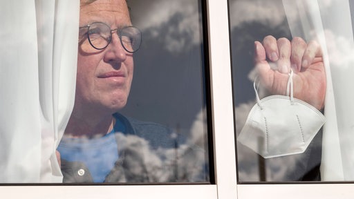 Ein Mann mit Maske in der Hand schaut sehnsüchtig aus seinem Fenster