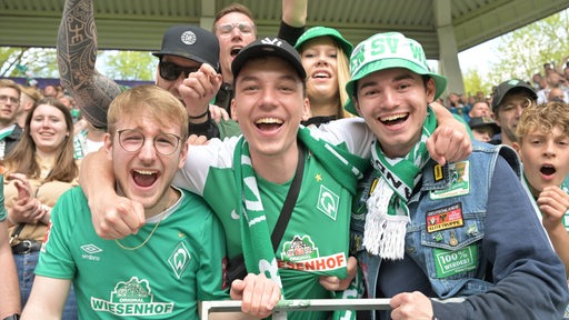 Werder-Fans stehen auf der Tribüne und jubeln strahlend in die Kamera.