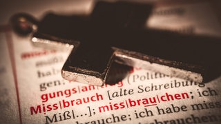 Ein Kreuz liegt auf einem Buchseite, auf der das Wort "Missbrauch" zu lesen ist