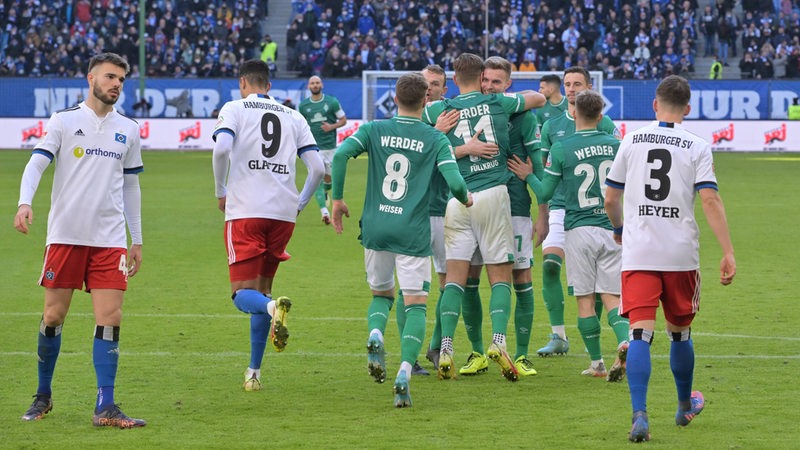 HSV-Spieler stehen enttäuscht auf dem Rasen, während Werder-Profi sich umarmen und das Tor bejubeln.