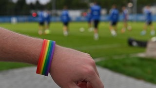 Zu sehen ist ein Arm mit einem Regenbogenarmband vor einem Fußballplatz-