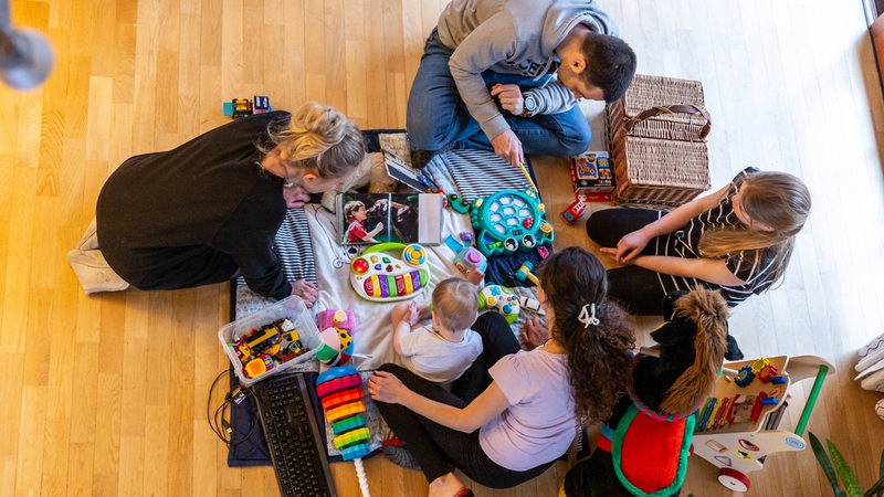 Vier Erwachsene sitzen mit einem Kleinkind auf einer Spieldecke. Um sie herum liegt Spielzeug verteilt.