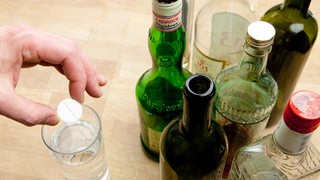 Ein Mann wirft eine Brausetablette in ein Glas Wasser, das neben leeren und halbvollen Flaschen Alkohol steht. 