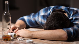 Ein Mann liegt mit dem Kopf auf einem Tisch, vor ihm eine fast leere Flasche Alkohol und Tablettten