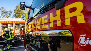 Blick auf einen Feuerwehrwagen. Im Hintergrund arbeiten zwei Feuerwehrmänner.