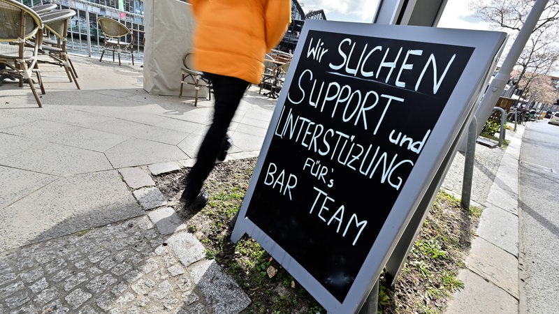 Eine Bar sucht mit einem Schild nach Unterstützung nach Personal. Ein Mensch geht an dem Schild vorbei. 