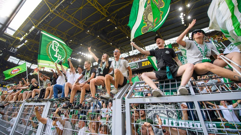 Werder-Fans sitzen glücklich und Fahnen schwenkend oben auf dem Zaun des Gästefanblocks im Dortmunder Westfalenstadion.