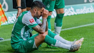 Werder-Spieler Leonardo Bittencourt sitzt auf dem Rasen und hält sich im Schmerz die Hände vors Gesicht.