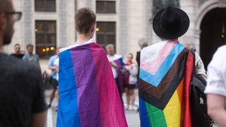 Bei einer Gedenkveranstaltung stehen zwei Menschen in transgender und Regenbogen-Flaggen gehüllt.