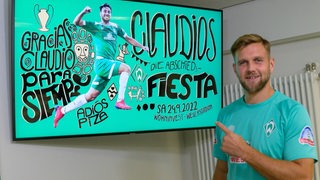 Werder-Spieler Niclas Füllkrug deutet lächelnd auf das Plakat zum Abschiedsspiel von Claudio Pizarro hin.