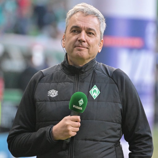 Werder-Stadionsprecher Arnd Zeigler hält lächelnd ein Mikrofon in der Hand.