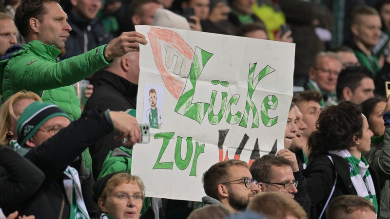 Ein Werder-Fan hält auf der Tribüne ein selbstgemaltes Schild hoch mit der Aufschrift "Lücke zur WM".