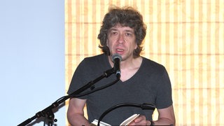 Ein Mann mit braun-grauem gelocktem Haar steht an einerm Rednerpult mit einem Buch in der Hand