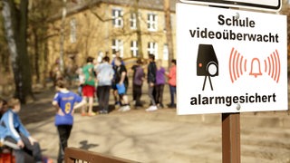 Auf einem Schild steht "Schule videoüberwacht – alarmgesichert". Im Hintergrund sind Kinder zu erkennen.