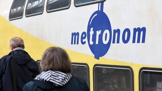 Mehrere Zuggäste schlendern an einem auf dem Gleis stehenden Metronomzug entlang.