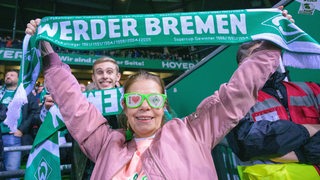 Ein weiblicher Werder-Fan mit "I love Werder"-Aufschrift auf der grünen Sonnenbrille hält im Stadion lächelnd einen Werder-Schal hoch.