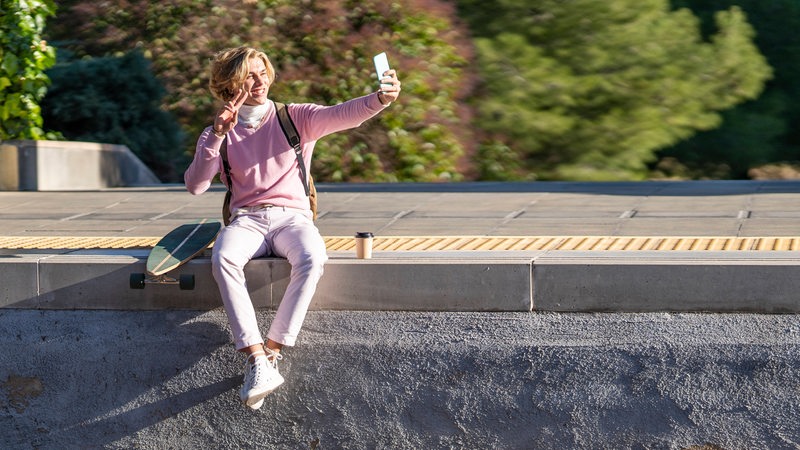 Ein junger Mann sitzt auf dem Bahnsteig und lässt die Beine ins Gleisbett baumeln. Davon macht er ein Selfie mit seinem Smartphone.