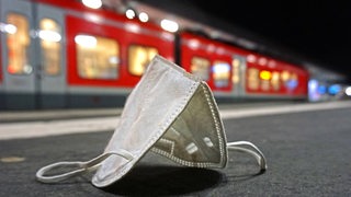 Eine weiße FFP2-Maske liegt auf einem Bahnsteig. Im Hintergrund ist ein Zug zu sehen.