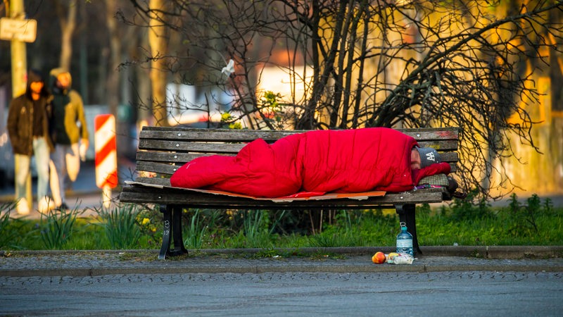 Ein Mann liegt in einem roten Schlafsack auf einer Parkbank.