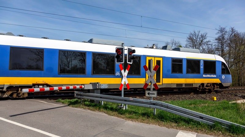 Ein blau-weiß-gelber Zug fährt über einen beschrankten Bahnübergang.
