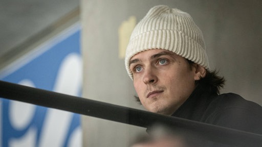 Der neue Eishockey-Spieler der Fischtown Pinguins sitzt auf der Tribüne mit einer weißen Wollmütze und schaut zu.