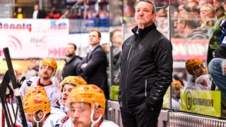 Pinguins-Coach Thomas Popiesch schaut mit den Händen in der Tasche konzentriert mit seinen Ersatzspielern auf das Eishockey-Spiel seines Teams.
