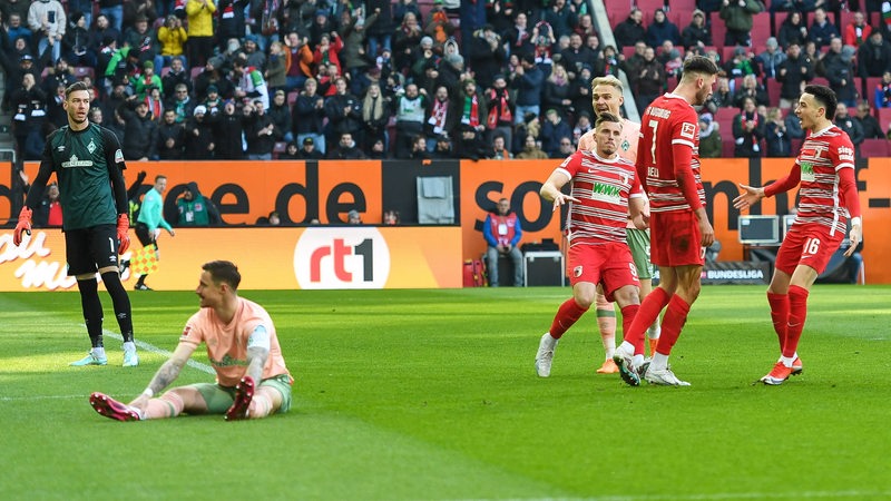 Werder-Kapitän Marco Friedl sitzt nach dem Gegentreffer frustriert auf dem Rasen, neben ihm jubeln Augsburger Spieler.