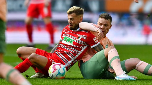 Werder-Spieler Niklas Schmidt verhakt sich sitzend im bissigen Zweikampf um den Ball mit Augsburgs Daniel Caligiuri.