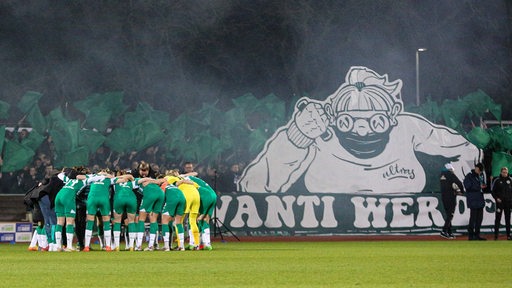 Werders Fußballerinnen bilden vor der Partie einen Motivationskreis auf dem Spielfeld, hinter ihnen einen großes Plakat der Werder-Ultras.