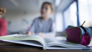 Eine junge Schülerin sitzt in der Klasse, im Vordergrund ein Schulbuch.